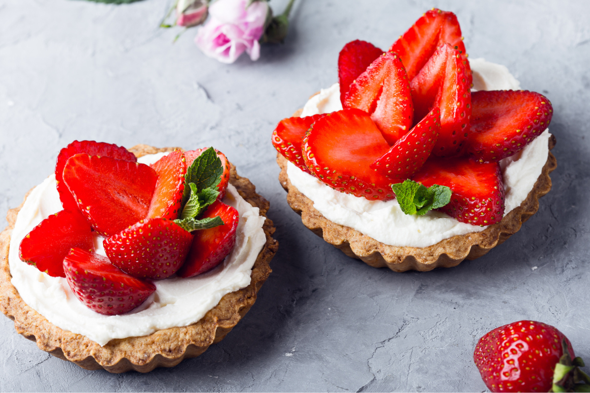 Cream cheese tartlets topped with fresh sliced strawberries and mint leaves on a textured gray surface.